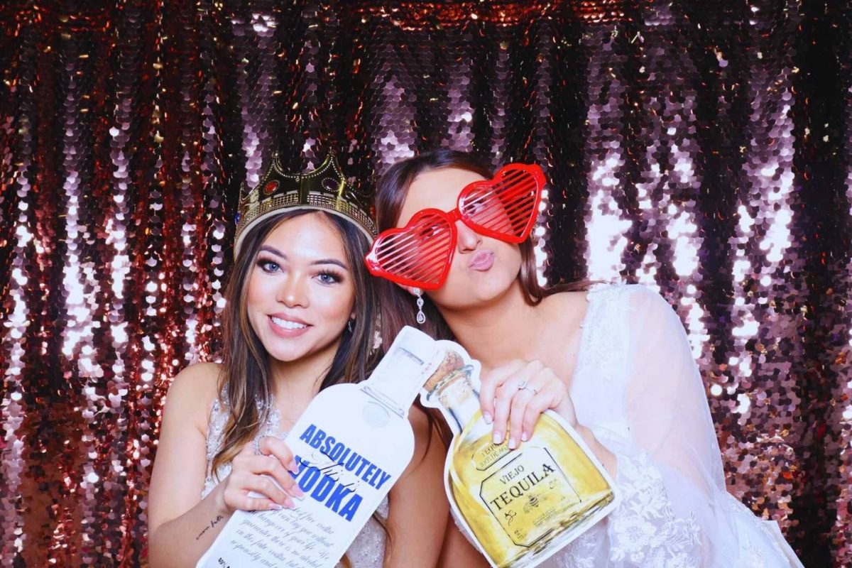 Two women in a wedding photo booth, one is the bride, both smiling and posing with fun props.