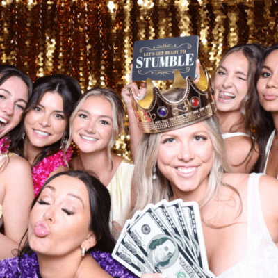 Bride and wedding guests posing with fun props in a photo booth with a gold sequin backdrop during a wedding reception